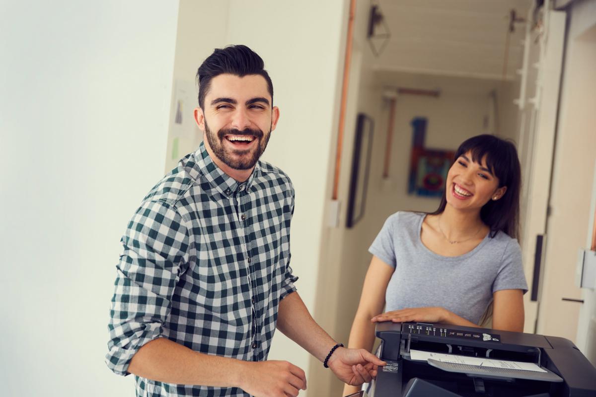 Two smiling coworkers collecting a printed document from an office copier.