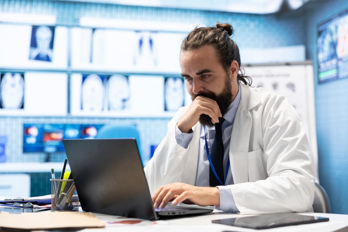 Doctor in a white coat reviewing patient imaging records on a laptop in a hospital office.