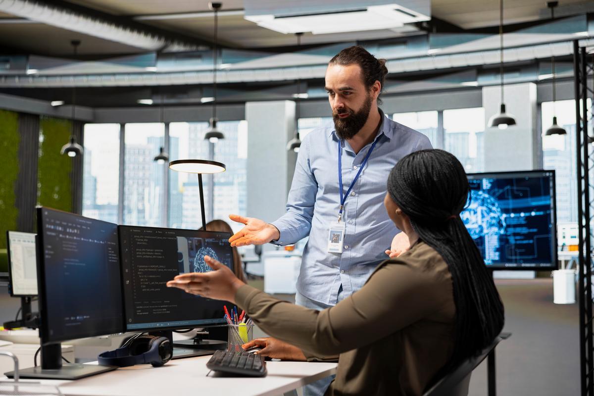 Two employees discussing code and data on desktop monitors in an open office.