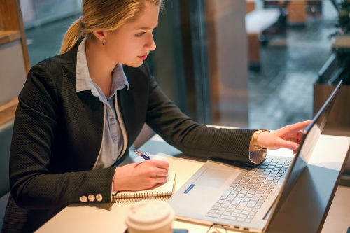 Woman concentrating and writing in notebook while using laptop